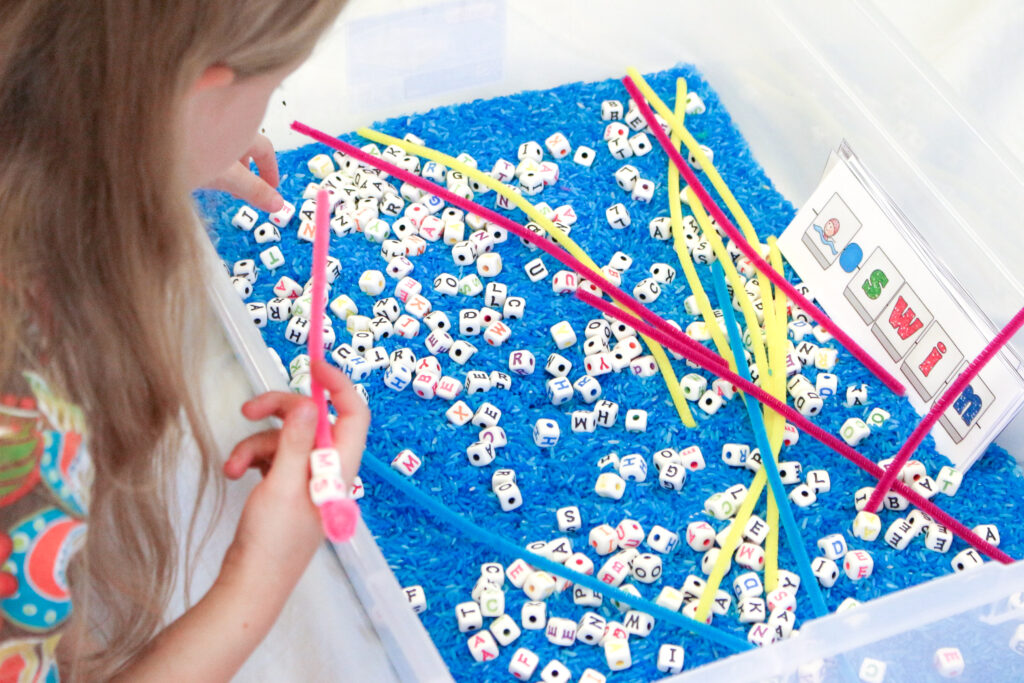 Image of young girl playing with alphabet beads in a sensory bin filled with blue rice and alphabet beads.